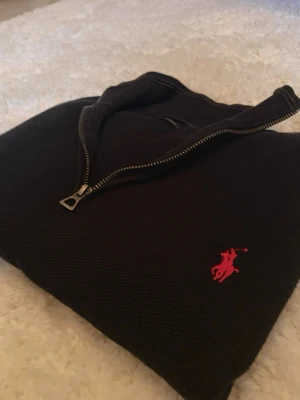 Svart stickad tröja Polo Ralph Lauren - Svart stickad tröja från Polo Ralph Lauren med halv dragkedja och röd broderad logga på bröstet. Tröjan har hög krage och ribbade muddar vid ärmslut och nederkant. Perfekt för lager på lager och klassisk stil.