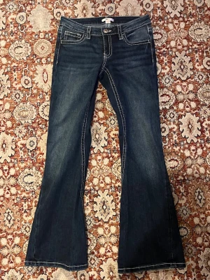 Bootcut jeans från Gina Tricot - Mörkblå bootcut jeans från Gina Tricot med vita kontrastsömmar och klassisk femficksdesign. Jeansen har hög midja och utsvängda ben, vilket ger en retroinspirerad look. Tillverkade i slitstark denim med snygga detaljer vid fickor och linning.