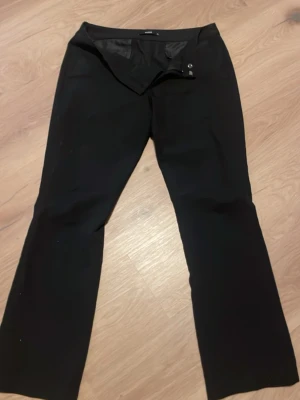 Svarta kostymbyxor med utsvängda ben - Svarta kostymbyxor med klassisk passform och utsvängda ben. Byxorna har enkel knäppning fram och är sydda i ett slätt tyg som ger en stilren look. Perfekta för dig som gillar en clean och tidlös stil.