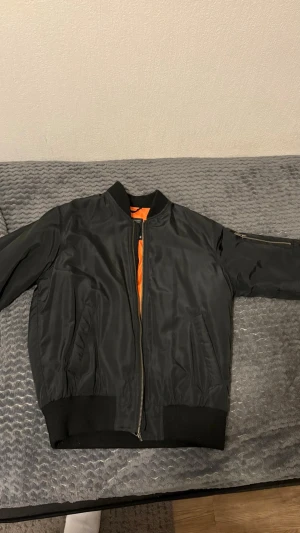 Svart bomberjacka med orange foder - Snygg svart bomberjacka från Topman med klassisk orange insida. Jackan har ribbade muddar, dragkedja framtill och fickor på sidorna. Materialet är glansigt och jackan har en ficka med dragkedja på ena ärmen. Perfekt för en cool streetstil.
