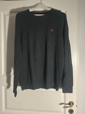 Mörkgrå stickad tröja från Morris - Mörkgrå stickad tröja med rund hals och diskret röd logga broderad på bröstet. Tröjan har långa ärmar och ribbade muddar vid ärmslut och nederkant. Perfekt för lager på lager och enkel att matcha med olika outfits.