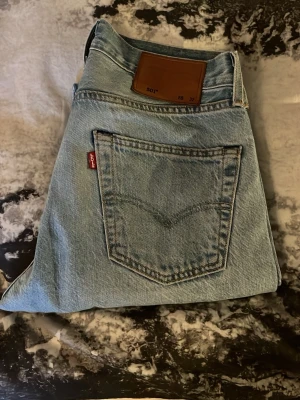 Levi's 501 ljusblå raka jeans - Klassiska Levi's 501 jeans i ljusblå denim med raka ben och femficksmodell. Jeansen har den ikoniska läderpatchen bak i midjan och röd Levi's-tagg på bakfickan. Tillverkade i 100% bomull för en autentisk vintagekänsla.