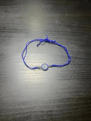 Blått armband med pärldetaljer - Snyggt armband med blå justerbar tråd och ett runt hänge dekorerat med små pärlor i blått, vitt och svart. Perfekt accessoar för att ge din outfit en färgklick och unik touch.