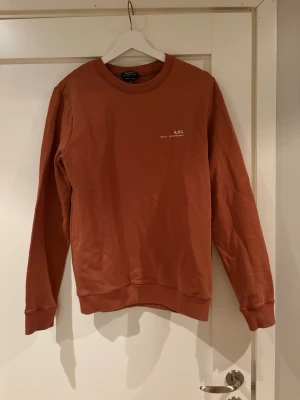 Rostbrun sweatshirt från A.P.C. - Säljer en stilren rostbrun sweatshirt från A.P.C. med rund halsringning och diskret vit logga på bröstet. Tröjan har långa ärmar, ribbade muddar och är gjord i mjuk bomull. Perfekt för en avslappnad och trendig look.