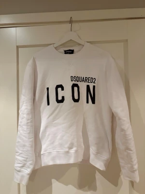 Vit sweatshirt DSQUARED2 ICON - Vit sweatshirt från DSQUARED2 med svart text 'ICON' och logga på bröstet. Tröjan har rund halsringning, långa ärmar och ribbade muddar. Perfekt för en trendig och avslappnad look.