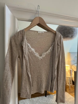 Beige kofta med spetsdetalj - Söt beige kofta med långa ärmar och v-ringning. Inbyggt linne i samma färg med vit spetsdetalj. Från Zara i stlk M men sitter som en S. Aldrig använd 