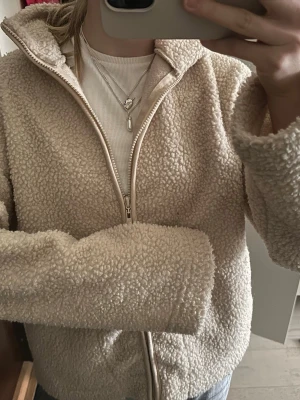 Beige teddyjacka från Amisu - Supermjuk teddyjacka i beige från Amisu. Jackan har dragkedja framtill, hög krage och är tillverkad i fluffigt teddy-material som ger en mysig och trendig look. 