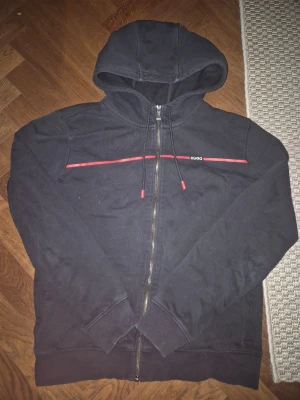 HUGO hoodie - Snygg svart hoodie från HUGO med dragkedja framtill och huva med dragsko. Röd detaljlinje över bröstet och diskret HUGO-logga. Tillverkad i mjukt bomullsmaterial som känns skönt mot huden. Perfekt för en avslappnad och stilren look.