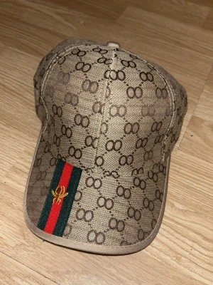 Beige keps med GG-mönster och rand - Säljer en beige keps från Gucci med det klassiska GG-monogrammet över hela kepsen. Framtill finns en grön och röd rand med guldfärgad brodyr. Justerbar rem baktill i samma tyg. Perfekt för dig som vill ha en lyxig och trendig accessoar. OBS fake