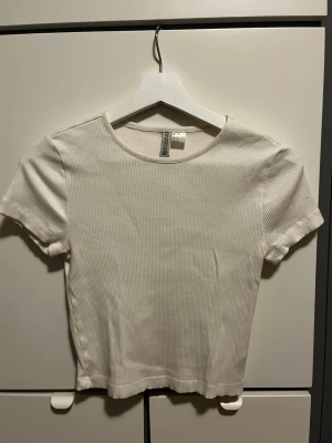 Vit ribbad t-shirt från Divided - En enkel och stilren vit ribbad t-shirt från Divided. Modellen är croppad med korta ärmar och rund halsringning. Perfekt att matcha med jeans eller kjol för en clean look.