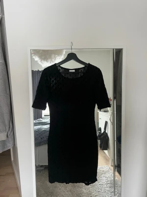 Svart prickig kortklänning - Snygg svart kortklänning med prickigt mönster och rund halsringning. Klänningen har korta ärmar och är tillverkad i ett lätt, transparent material upptill. Perfekt för dig som gillar en stilren look med en twist.