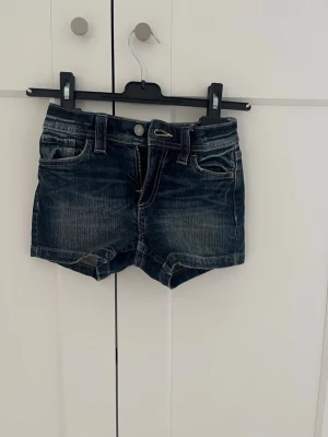 Mörkblå jeansshorts med fickor - Snygga mörkblå jeansshorts med klassisk femficksdesign och slitna detaljer. Storlek 134/xxs.Shortsen har knapp och dragkedja framtill samt bakfickor med lock och knapp. Perfekta för varma dagar och enkel att matcha med allt.Säljer pga för små.
