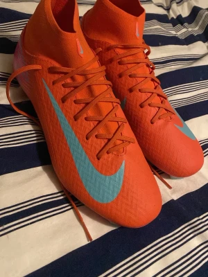 Nike orange fotbollsskor med blå swoosh - Snygga fotbollsskor från Nike i en stark orange färg med ljusblå swoosh på sidan. Skorna har högt skaft, snörning och är tillverkade i ett lätt syntetmaterial som ger bra bollkontroll. Perfekta för dig som vill sticka ut på planen.