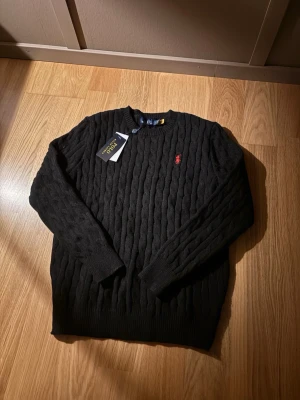 Svart kabelstickad tröja från Polo Ralph Lauren - Kabelstickad svart tröja från Polo Ralph Lauren med klassisk röd broderad logga på bröstet. Tröjan har rund halsringning och ribbade muddar vid ärmslut och nederkant. Är ganska liten i storleken och passar både S och M med prislapp!
