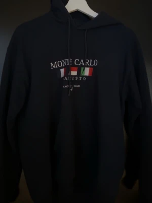 Svart hoodie Monte Carlo Yacht Club - Svart hoodie från Monte Carlo Yacht Club med broderad text och flaggor på bröstet. Klassisk modell med huva och dragsnören. Mjuk och skön bomullsmix, perfekt för chill dagar. Långärmad och stilren design.