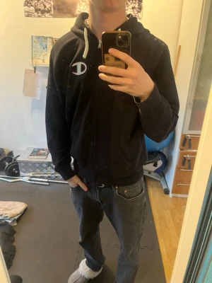 Svart zip hoodie från Champion - Säljer en svart hoodie från Champion med dragkedja och vit snörning i huvan. Stor Champion-logga på bröstet och fickor framtill. Skön och avslappnad passform, perfekt till jeans.