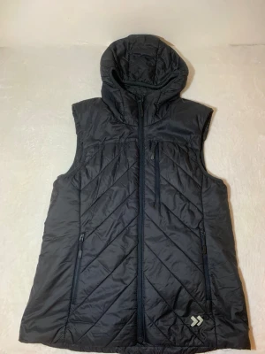 Svart dunväst med huva - Snygg svart dunväst med huva och dragkedja framtill. Västen har tre fickor med dragkedja och ett diskret märke nertill. Snygga diagonala sömmar ger en sportig look. Perfekt för lager-på-lager och funkar året runt.