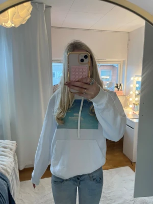 Vit hoodie med blått tryck - Säljer en vit hoodie med huva och dragsnören. På bröstet finns ett stort tryck med ett blått himmel- och molnmotiv. Hoodien har en klassisk känguruficka framtill och är i mjukt material, perfekt för chill dagar.