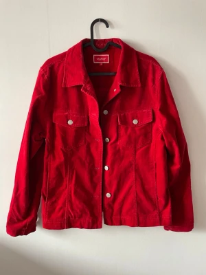 Röd jacka  - Snygg röd jacka med klassisk krage,silverfärgade knappar och två bröstfickor och två fickor längre ner. Jackan har raka ärmar och materialet är i ett sammetsliknade-material.