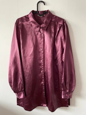 Lila satinskjorta med knappar - Lila skjorta i glansigt satinmaterial med klassisk krage och knäppning framtill. Skjortan har långa ärmar med manschett och en loose passform.