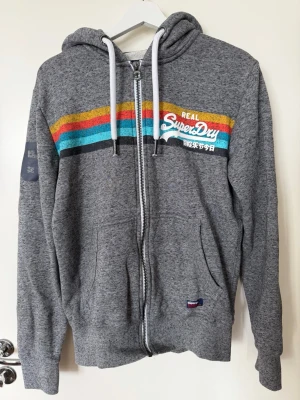 Grå Superdry hoodie med dragkedja - Grå hoodie från Superdry med dragkedja, huva och vita snören. Färgglada ränder i gult, rött och blått över bröstet samt Superdry-logga tryckt på vänster sida. Mjuk, fleecefodrad insida och två fickor framtill. Perfekt för en avslappnad streetwear-look.