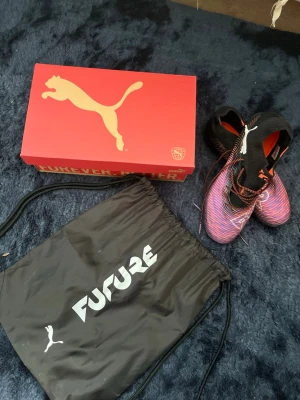 Puma Future Elit fotbollsskor svart/lila - Säljer ett par Puma Future Elit fotbollsskor i svart och lila med vita detaljer. Skorna har en modern design med snörning och låg profil, samt en ovandel i syntetmaterial för bra bollkontroll. Levereras med originalpåse och låda. Perfekta för dig som vill sticka ut på planen. Pris kan diskuteras.