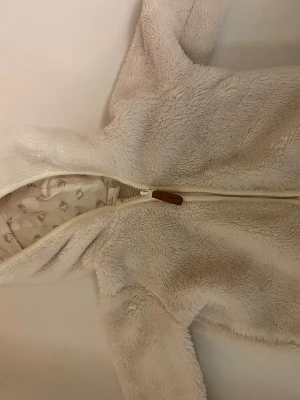 Vit teddyjacka med huva - Supermjuk vit teddyjacka med dragkedja och huva. Jackan har ett gulligt foder med igelkottsmönster och är tillverkad i fluffigt teddy-material som håller dig varm. Perfekt för dig som gillar mysiga och stilrena plagg.