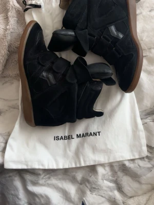 Svarta Isabel Marant sneakers - Snygga svarta sneakers från Isabel Marant med högt skaft och kardborreband. Skorna har en cool design i mocka och läderdetaljer samt brun sula. Perfekta för dig som vill ha en trendig och unik look.
