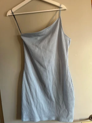 Ljusblå randig oneshoulder klänning - Snygg ljusblå kortklänning med tunna vita ränder och oneshoulder-design. Klänningen har smala axelband och är tillverkad i ett mjukt, stretchigt material som sitter bekvämt. Perfekt för dig som gillar en stilren och trendig look.