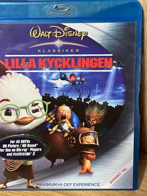 Lilla Kycklingen Blu-ray Disney - Blu-ray-film av Walt Disney, Lilla Kycklingen, i klassiker-serien. Omslaget är blått med färgglad tecknad framsida och svensk text. Innehåller extramaterial som borttagna scener och karaoke. Svenskt tal och flera språkval.