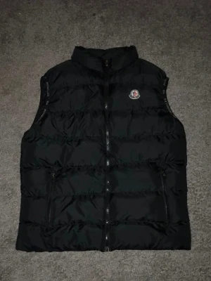Svart väst från Moncler - Säljer nu en riktigt fet väst från Moncler. Västen är inte äkta, men eftersom att den har det en riktig har så kommer ingen fatta. Även fast storleken är L så passar den M också. Västen är nästan aldrig använd. Loggan ser perfekt ut, den har bra dragkedjior och värmer där den täcker. Perfekt inför sommaren och därmed väldigt enkel att styla. Hör av dig vid frågor!