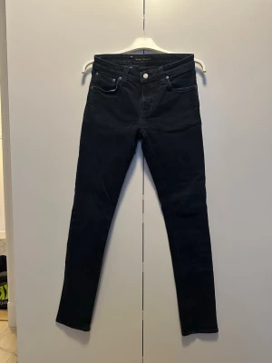 Svarta slim jeans från Nudie Jeans - Snygga svarta slim jeans från Nudie Jeans med klassisk femficksmodell och smal passform. Jeansen har normal midja och är tillverkade i stretchigt denimtyg för extra komfort. Perfekta för en stilren och modern look.
