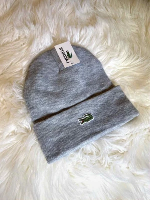Grå mössa från Lacoste - Snygg grå ribbstickad mössa från Lacoste med klassiska gröna krokodilloggan framtill. Mössan har uppvikt kant och är tillverkad i mjukt material som håller dig varm. Perfekt accessoar för kyliga dagar.