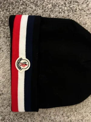 Svart Moncler mössa med ränder - Snygg svart mössa från Moncler med breda ränder i rött, vitt och blått vid uppviket. Klassisk Moncler-logga framtill. Tillverkad i mjukt material som sitter skönt på huvudet. Perfekt accessoar för dig som vill ha en stilren och trendig look.
