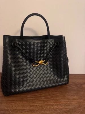 Svart flätad handväska med gulddetalj - Snygg svart handväska i flätat läder med guldfärgad metallknut framtill. Väskan har dubbla handtag, innerfack med dragkedja och stilrena detaljer. Perfekt för dig som gillar klassisk men trendig design och vill ha plats för allt du behöver.