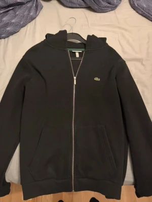 Svart hoodie med dragkedja från Lacoste - Svart hoodie från Lacoste med hel dragkedja framtill och klassisk krokodillogga på bröstet. Hoodien har två fickor fram, ribbade muddar och huva. Perfekt för en chill och stilren look.