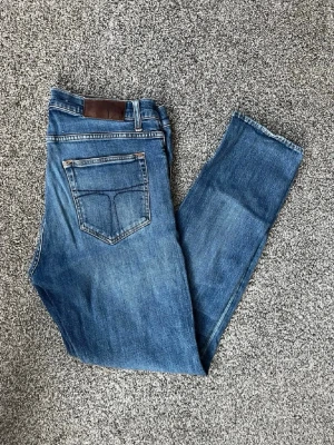 Blå jeans från tiger of sweden - Snygga blå jeans med rak passform och klassisk femficksdesign. Jeansen har en mörk läderpatch bak i midjan och slitna detaljer framtill för en avslappnad look. Tillverkade i denim med kontrastsömmar och normal midja.