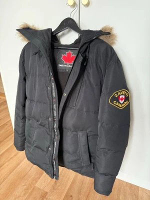 Svart dunjacka från Zavetti Canada - Snygg svart dunjacka från Zavetti Canada med fuskpäls. Jackan har patch med logga på ärmen, dragkedja och knappar framtill samt två stora fickor. Perfekt för kalla vinterdagar och riktigt varm.