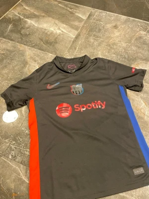 Svart FC Barcelona Nike matchtröja - Snygg svart FC Barcelona matchtröja från Nike med röd och blå detalj på sidorna. Tröjan har klubbmärke, Nike-logga och Spotify som sponsor framtill. Tillverkad i lätt och ventilerande material, perfekt för fotboll eller träning.