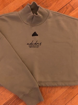 Olivgrön cropped hoodie med tryck - Snygg olivgrön cropped hoodie från Adidas med hög krage och svart tryck på bröstet. Mjuk bomullskänsla och ribbade muddar vid ärmsluten. Perfekt för en avslappnad streetwear-look.