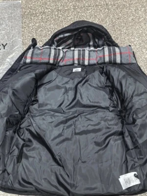 Svart pufferjacka från Burberry - Snygg svart pufferjacka från Burberry med klassiskt rutigt foder i grått, rött och vitt. Jackan har huva, dragkedja och tryckknappar framtill samt Burberry-logga på ärmen. Perfekt för kalla vinterdagar och riktigt trendig stil.