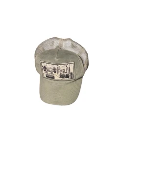 Beige truckerkeps Emilio Pendenza - Snygg beige truckerkeps från Emilio Pendenza med mesh på sidorna och baksidan. Framtill finns ett tryck med ett svartvitt gatufoto. Kepsen har böjd skärm och justerbar passform. Perfekt för en avslappnad streetwear-look.