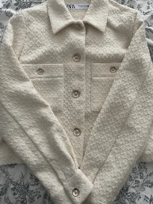 Krämvit overshirt från Zara - Stilren krämvit overshirt från Zara med strukturerat tyg och dekorativa guldiga knappar. Två stora bröstfickor med lock och klassisk krage ger en trendig look. Perfekt att bära öppen eller stängd för en cool lager-på-lager-stil.