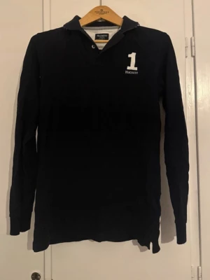 Svart långärmad skjorta Hackett - Snygg svart långärmad skjorta från Hackett med vit broderad siffra 1 på bröstet och ärmen. Klassisk krage och knappslå framtill.