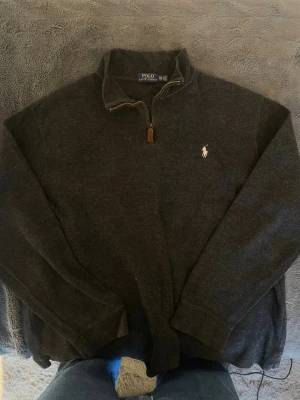 Mörkgrå halvzip från Polo Ralph Lauren - Mörkgrå tröja med halv dragkedja och ståkrage från Polo Ralph Lauren. Klassisk broderad logga i vitt på bröstet och mjukt material som känns skönt mot huden. Perfekt för lager på lager och enkel att matcha med jeans eller chinos.