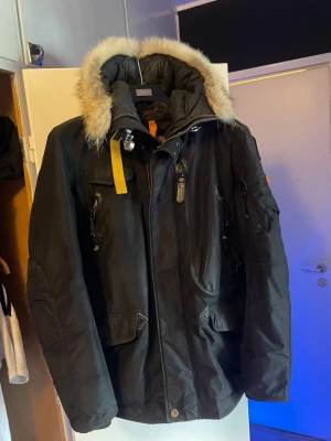 Svart parkasjacka från Parajumpers - Säljer en svart parkasjacka från Parajumpers med avtagbar huva och fluffig fuskpäls. Jackan har flera fickor, dragkedja och tryckknappar framtill samt den klassiska gula remmen vid kragen. Perfekt för kalla dagar och riktigt snygg streetstil.