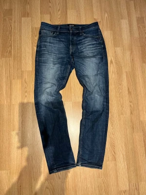 Mörkblå raka jeans med slitningar - Säljer ett par mörkblå jeans med raka ben och snygga slitningar framtill. Klassisk femficksmodell med knappgylf och kontrastsömmar. Jeansen har en läderpatch bak i midjan och är tillverkade i denim. Perfekta för en avslappnad och trendig look.