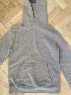 Grå basic hoodie med magficka - En enkel och stilren grå hoodie med stor magficka och luva . Hoodien har ribbade muddar vid ärmslut och nederkant. Perfekt för en avslappnad look och passar till det mesta. Materialet känns mjukt och bekvämt, troligen bomullsblandning.