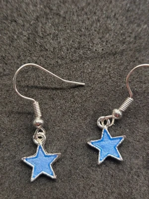 Blå lysande stjärnörhängen i rostfritt stål  - Säljer ett par coola örhängen med blå stjärnformade hängen som lyser i mörkret. Stjärnorna är gjorda i silverfärgade metalldetaljer och har krokar i rostfritt stål för enkel användning. Perfekta för dig som vill sticka ut med en unik accessoar!