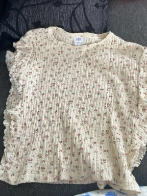 Blommig volangtop från Zara - Supersöt blommig topp från Zara i ribbad bomull med volangdetaljer längs ärmarna. Toppen är ljusbeige med små rosa och gröna blommor, har rund hals och är kortärmad. Perfekt för dig som gillar romantisk och trendig stil!